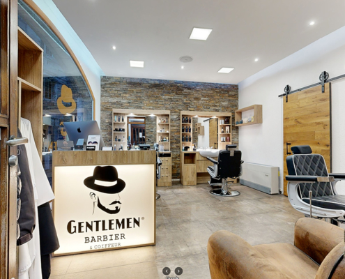 L'image présente l'intérieur élégant de "Gentlemen Barbier & Coiffeur" à Genève, mettant en vedette des fauteuils de barbier noirs, des murs en pierre, et un sol en bois, avec le logo distinctif du salon illuminé en premier plan.