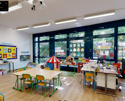 La photo montre une salle de classe lumineuse et colorée de "The British School of Geneva", avec des tables d'élèves, des chaises colorées, des affiches éducatives sur les murs et une zone de jeu pour enfants.