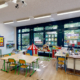 La photo montre une salle de classe lumineuse et colorée de "The British School of Geneva", avec des tables d'élèves, des chaises colorées, des affiches éducatives sur les murs et une zone de jeu pour enfants.