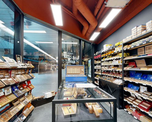 Cette photo montre l'intérieur du magasin de cigares, The Cigar Market, éclairé et organisé, avec des étagères remplies de boîtes de cigares et une vitrine centrale.
