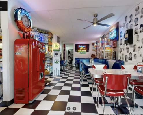 Cette image présente l'intérieur du Happy Days Diner dans un style américain rétro : sièges en vinyle rouge, sol damier noir et blanc, décoré de néons et de souvenirs vintage comme une pompe à essence Coca-Cola et des plaques d'immatriculation. L'ambiance évoque les années 50.