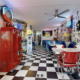 Cette image présente l'intérieur du Happy Days Diner dans un style américain rétro : sièges en vinyle rouge, sol damier noir et blanc, décoré de néons et de souvenirs vintage comme une pompe à essence Coca-Cola et des plaques d'immatriculation. L'ambiance évoque les années 50.