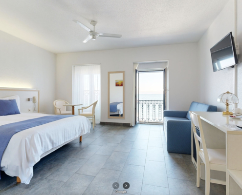 La photo présente la chambre "Business Grand Lit" de l'Hôtel du Port. Elle est éclairée et offre un espace généreux avec un lit double agrémenté de linge de lit blanc et d'une couverture bleue. La chambre dispose d'une porte-fenêtre qui s'ouvre sur un balcon qui donne sur le lac.