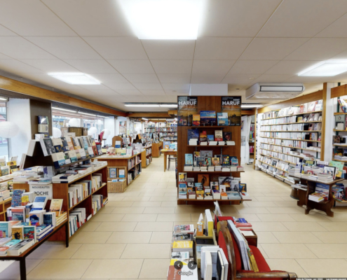 L'image montre l'intérieur d'une librairie spacieuse et bien éclairée. Des étagères remplies de livres s'alignent sur les murs et des présentoirs centraux offrent des sélections de nouveautés et de recommandations.