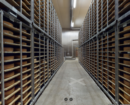 La photo montre l'intérieur de Vacherin Fribourgeois SA, où des centaines de meules de fromage sont soigneusement rangées sur des étagères métalliques. Ces étagères s'étendent sur toute la hauteur de la pièce, formant un couloir central par lequel on peut marcher.