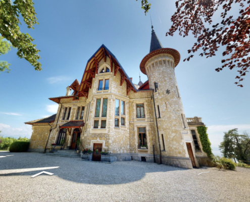 Cette image montre la façade extérieure d'une grande villa (château) avec vue sur le lac. Cette photo est prise depuis le jardin et donne sur l'entrée de la villa.