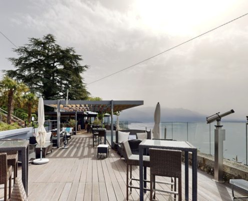 Cette image montre la terrasse spacieuse et élégante du bar "Le Deck", qui s'ouvre sur une vue imprenable. Avec son sol en bois, ses tables en osier confortables et ses chaises assorties, l'endroit invite à la détente et au plaisir des sens. Des télescopes sont installés à l'avant, suggérant que les clients peuvent profiter de vues rapprochées sur le lac Léman et les montagnes au loin.
