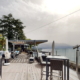 Cette image montre la terrasse spacieuse et élégante du bar "Le Deck", qui s'ouvre sur une vue imprenable. Avec son sol en bois, ses tables en osier confortables et ses chaises assorties, l'endroit invite à la détente et au plaisir des sens. Des télescopes sont installés à l'avant, suggérant que les clients peuvent profiter de vues rapprochées sur le lac Léman et les montagnes au loin.