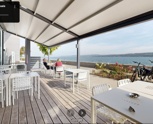L'image montre une terrasse spacieuse et bien aménagée, avec des tables blanches et des chaises assorties sous un auvent élégant. La terrasse offre une vue imprenable sur le lac de Neuchâtel.