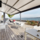 L'image montre une terrasse spacieuse et bien aménagée, avec des tables blanches et des chaises assorties sous un auvent élégant. La terrasse offre une vue imprenable sur le lac de Neuchâtel.