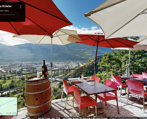 Cette image montre une terrasse accueillante avec mobilier rouge, sous parasols blancs et rouges, dominant Sion. Barrique en bois, bouteille de vin posée, invitation à la détente avec vue panoramique sur la vallée du Valais.