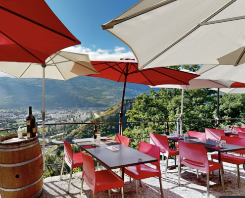 Cette image montre une terrasse accueillante avec mobilier rouge, sous parasols blancs et rouges, dominant Sion. Barrique en bois, bouteille de vin posée, invitation à la détente avec vue panoramique sur la vallée du Valais.