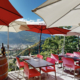 Cette image montre une terrasse accueillante avec mobilier rouge, sous parasols blancs et rouges, dominant Sion. Barrique en bois, bouteille de vin posée, invitation à la détente avec vue panoramique sur la vallée du Valais.