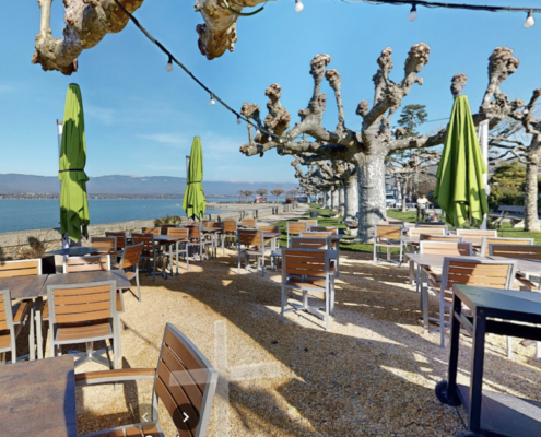 Cette image montre la terrasse du Café du Quai offre une ambiance relaxante avec des tables en bois, des parasols verts, surplombant le lac Léman et encadrée par des arbres sculpturaux.