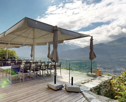 Cette image montre la terrasse élégante du restaurant "Les Mazots du Clos du Château", offrant une vue imprenable sur les vignes et la vallée de Sion.